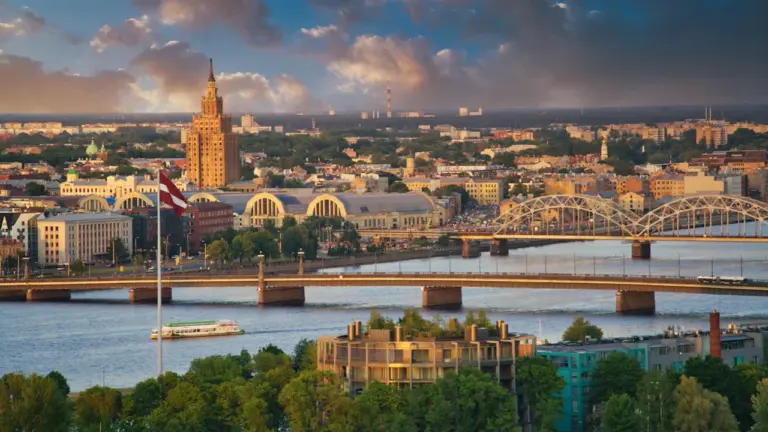 Panorāmas skats uz Rīgas vēsturisko centru un Daugavu ar Latvijas karogu priekšplānā.