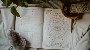 Atvērta dienasgrāmata ar astroloģiskiem simboliem, kristālu un augu uz mīkstas tekstila virsmas.