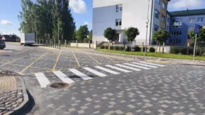 Gājēju pāreja uz bruģētas ielas pie moderna daudzstāvu ēkas saulainā dienā.