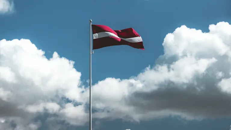 Sarkanbaltsarkanais Latvijas valsts karogs plīvo vējā pretī zilām debesīm ar baltiem mākoņiem.