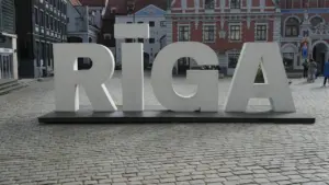 Liels uzraksts RĪGA uz bruģētas ielas vēsturiskajā pilsētas centrā saulainā dienā.