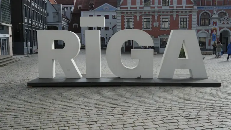 Liels uzraksts RĪGA uz bruģētas ielas vēsturiskajā pilsētas centrā saulainā dienā.