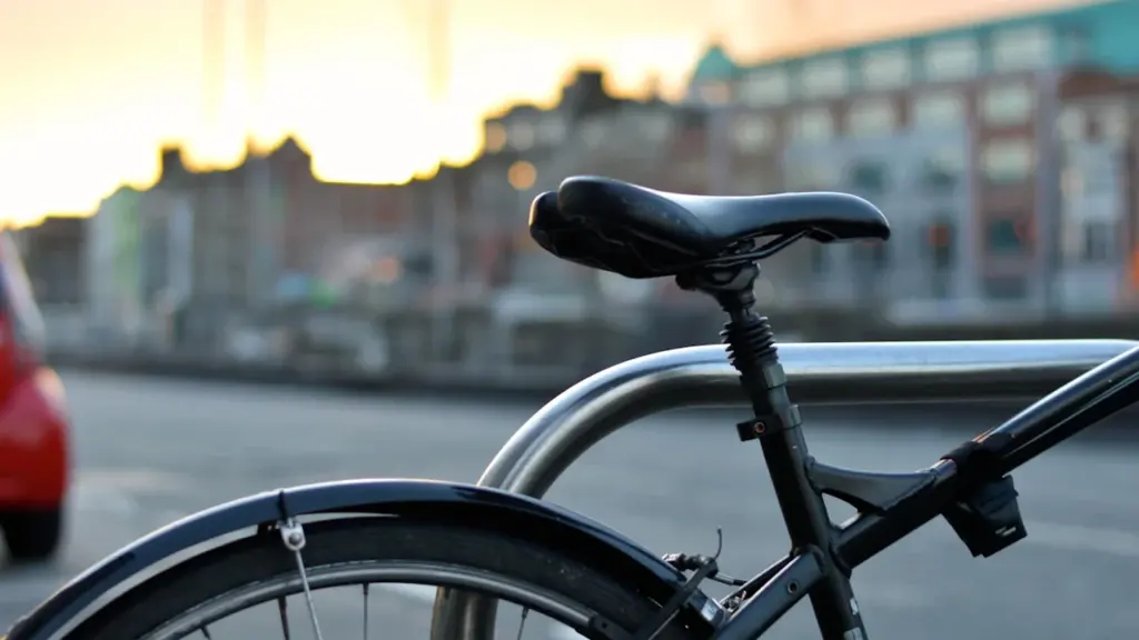 Pilsētas velosipēds novietots pie velostatīva pilsētas ielas malā saulrieta laikā.