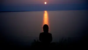 Cilvēka siluets meditē pie ezera saulrietā, simbolizējot garīgu mieru un klusumu.