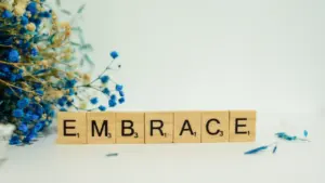 Koka klucīši ar vārdu 'embrace' un zilas pavasara puķes uz galda.