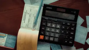 Kalkulators uz koka galda ar čupām papīra rēķinu un finanšu dokumentiem.