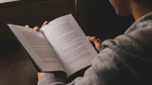 Cilvēks lasa atvērtu grāmatu ar tekstu, kas atgādina dzejas lasīšanu bibliotēkas vidē.