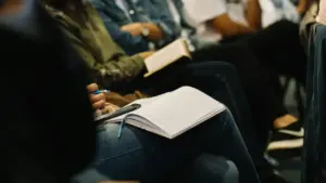 Cilvēki sēž auditorijā un tur klēpī atvērtas piezīmju grāmatiņas, gatavojoties pierakstīt svarīgu informāciju.